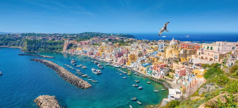 Panoramic view of beautiful Procida in sunny summer day. Colorful houses, cafes and restaurants, fishing boats and yachts in Marina Corricella, clear blue sky and azure sea in Procida Island, Italy.